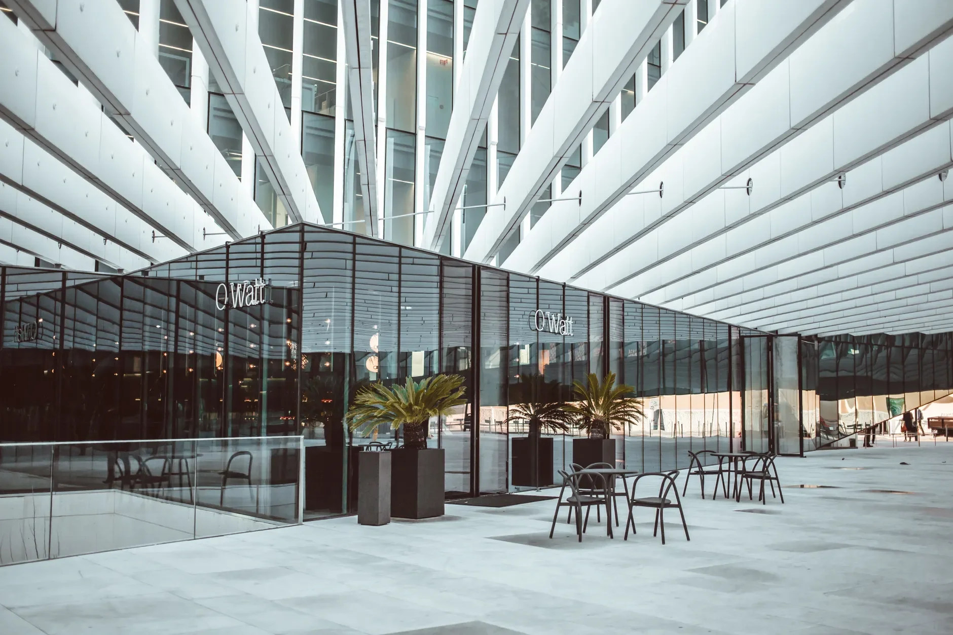 Gläserne Gewerbefassade mit Sitzplätzen und Pflanzkübeln unter einer weißen, geometrisch gegliederten Überdachung.